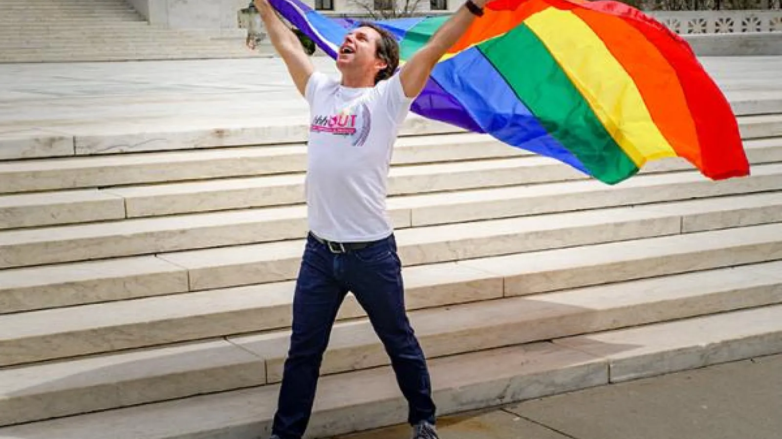 A person holding up the LGBT flag in front of government building stairs
