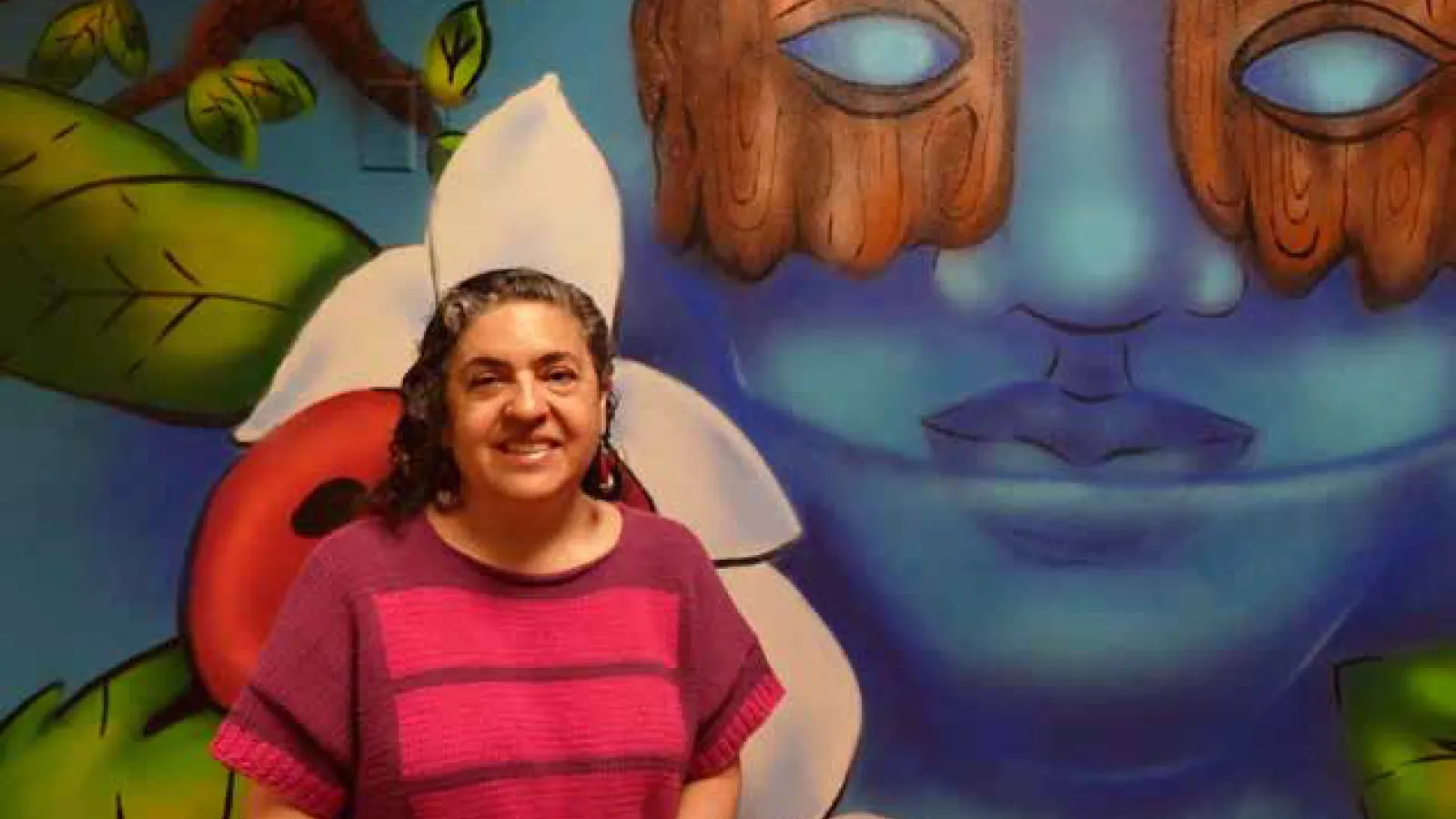 Guisela Latorre sitting in front of the mural in the WGSS department suite, 286 University Hall
