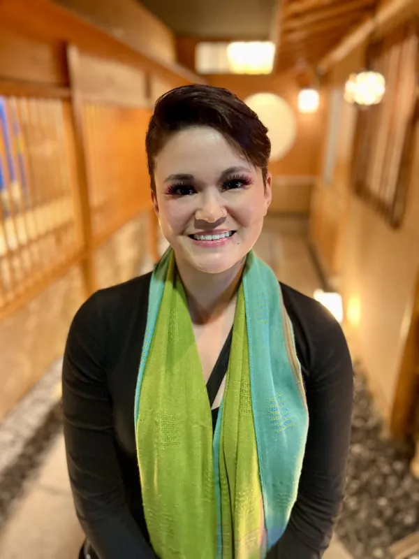 Photograph of Corinne Sugino smiling towards the camera while wearing a black shirt and green scarf