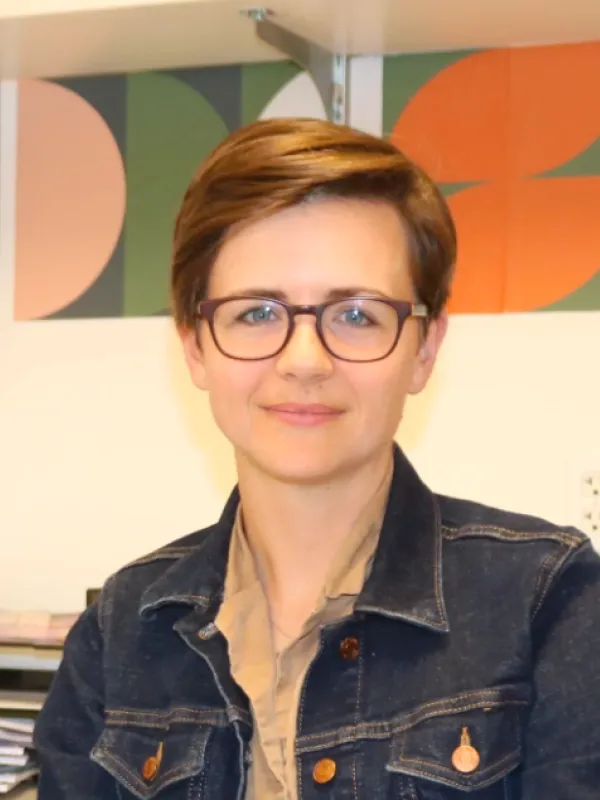 Person with short brown hair and glasses, jean jacket and brown shirt sitting facing the camera.